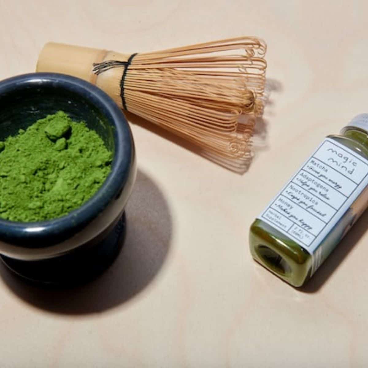 Green tea powder in a small black bowl with a mini matcha bottle on the side and a brown manual food mixer