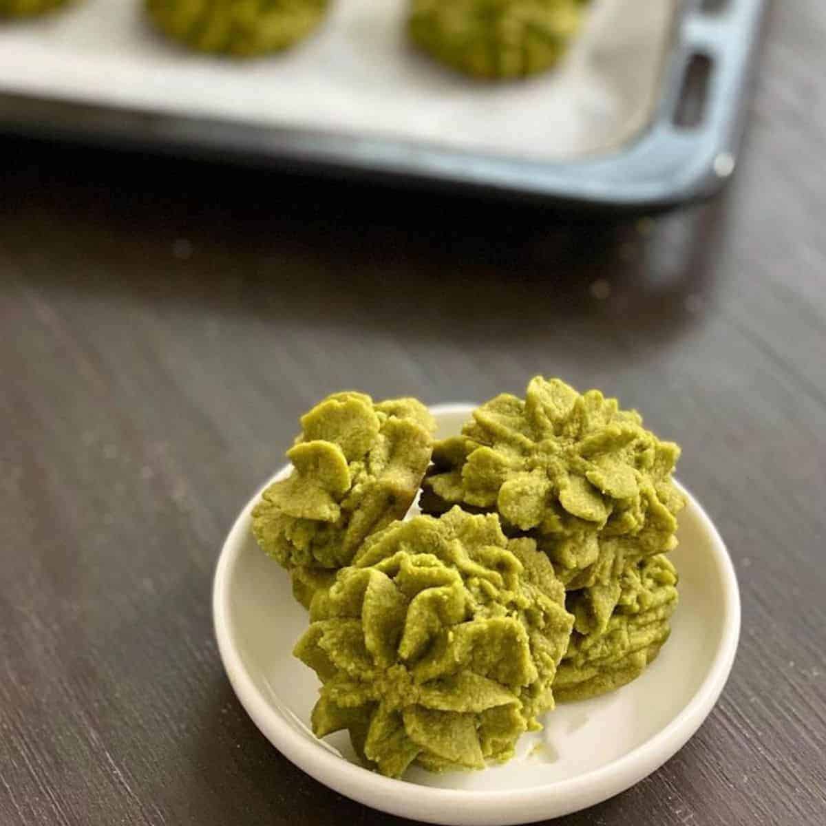 Mini green tea biscuits with artistic design in a small white plate with a blurry tray of cookies in the background