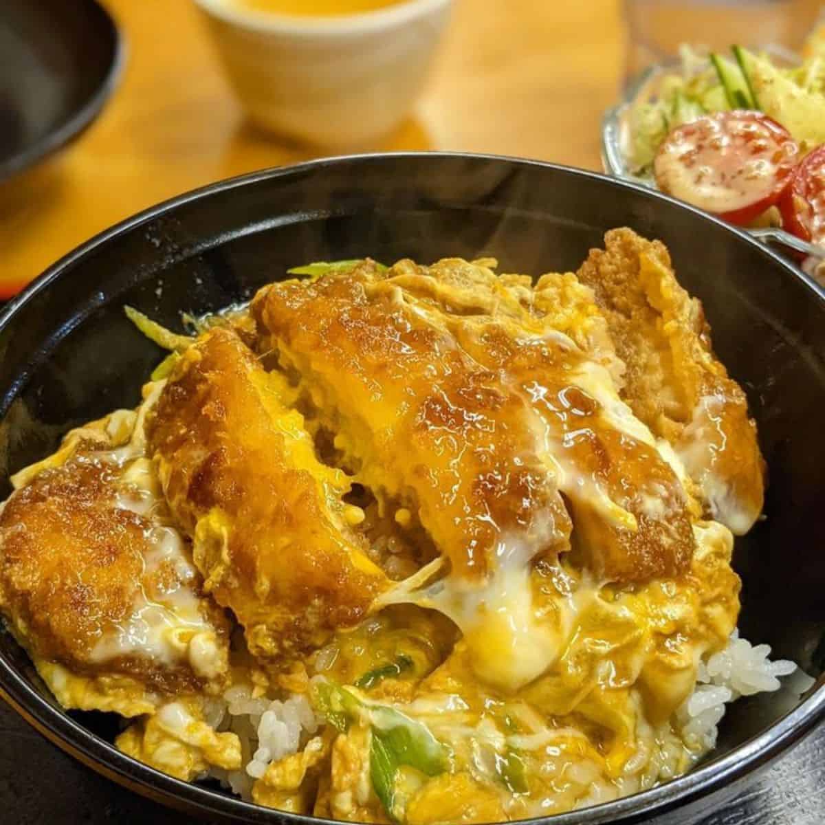 Katsudon in a black bowl with vegetable salad on another bowl