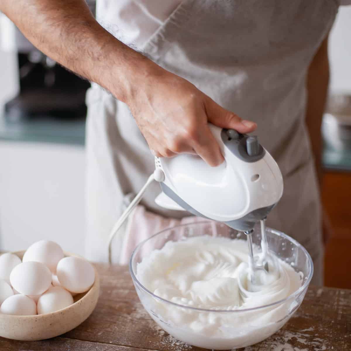 A baker using a hand mixer on a bowl of white cream with other ingredients on the table