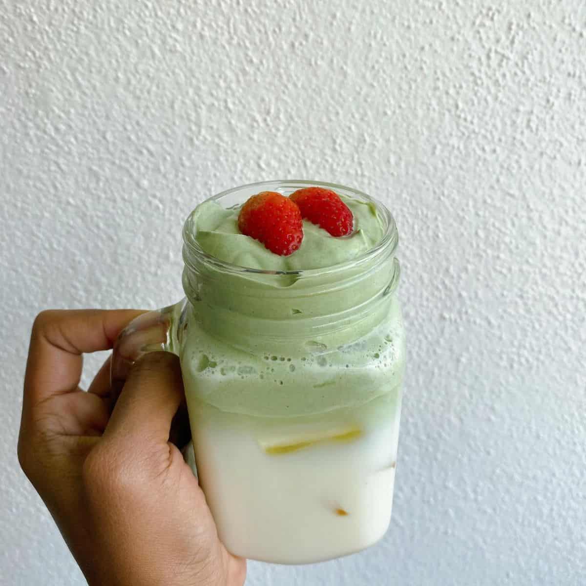 A hand holding a tempting Dalgona Matcha drink with slices of fresh strawberries on top.