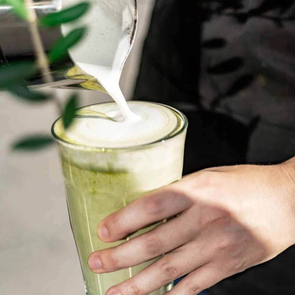 A person pouring out milk in the glass of a green tea beverage