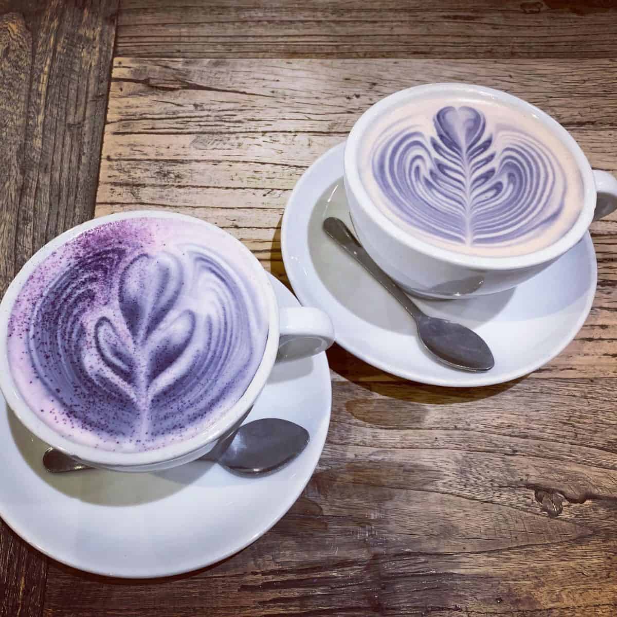 Two white ceramic cups filled with Lavender Milk Tea with saucers and spoons in a woody table