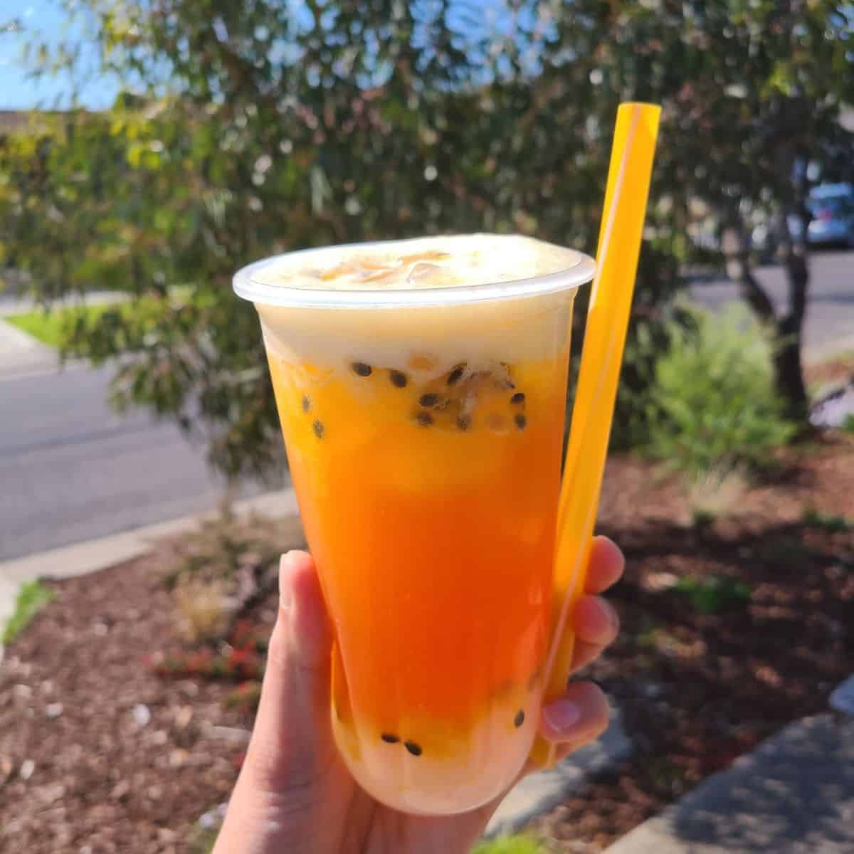  A hand holding an orange refreshment in a plastic cup with yellow straw on the roadside.