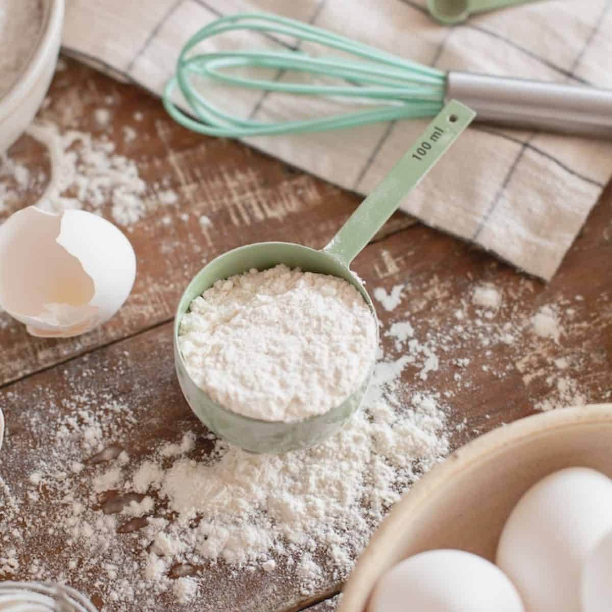 White powder in a measuring cup with eggs on the side and other kitchen tools